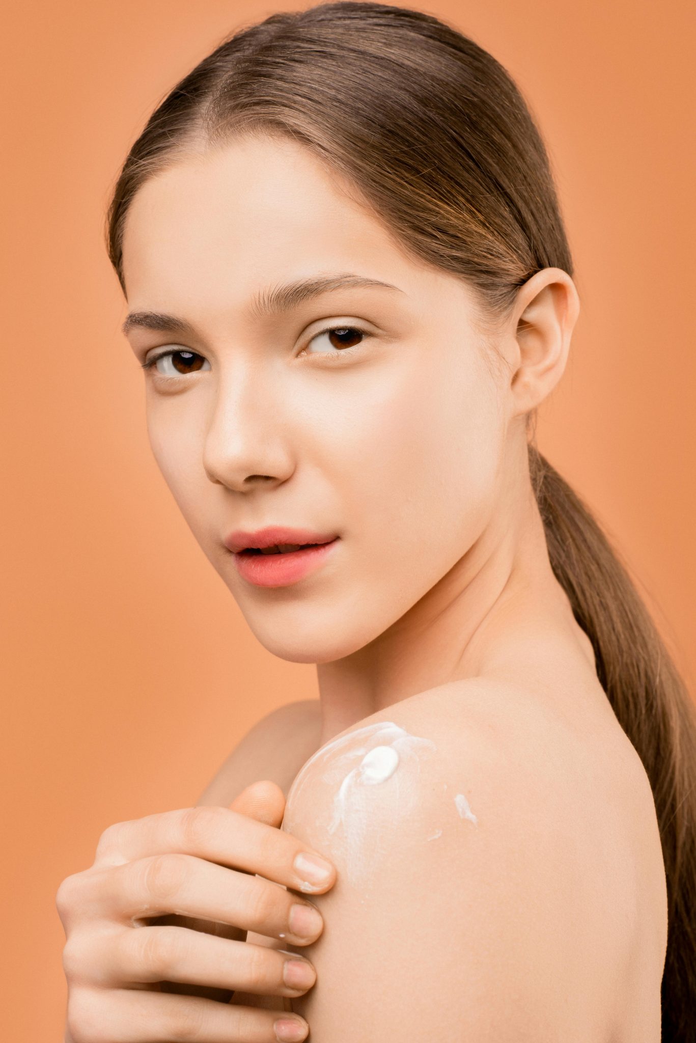 Young woman applies moisturizer on shoulder, set against a peach background, highlighting smooth skin care.